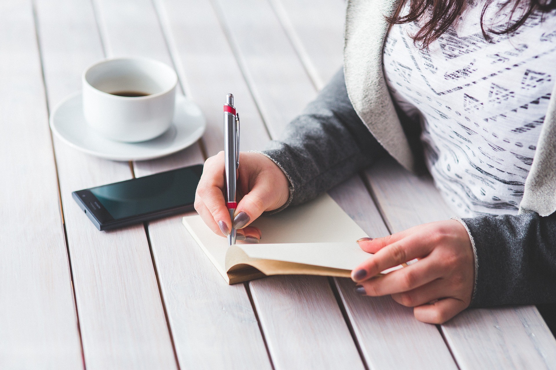 Woman sitting at a white table writing in a notebook with a cup of coffee and a mobile phone beside her.